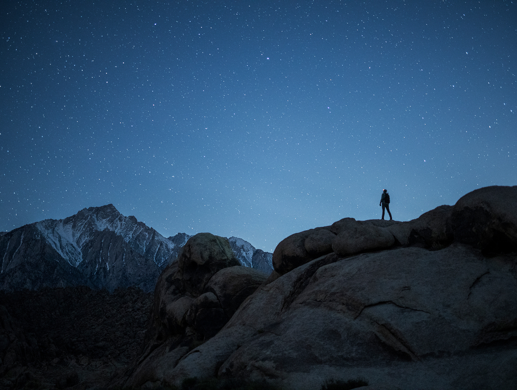 Far away nighttime shot of man on mountains surrounded by a star-filled sky.