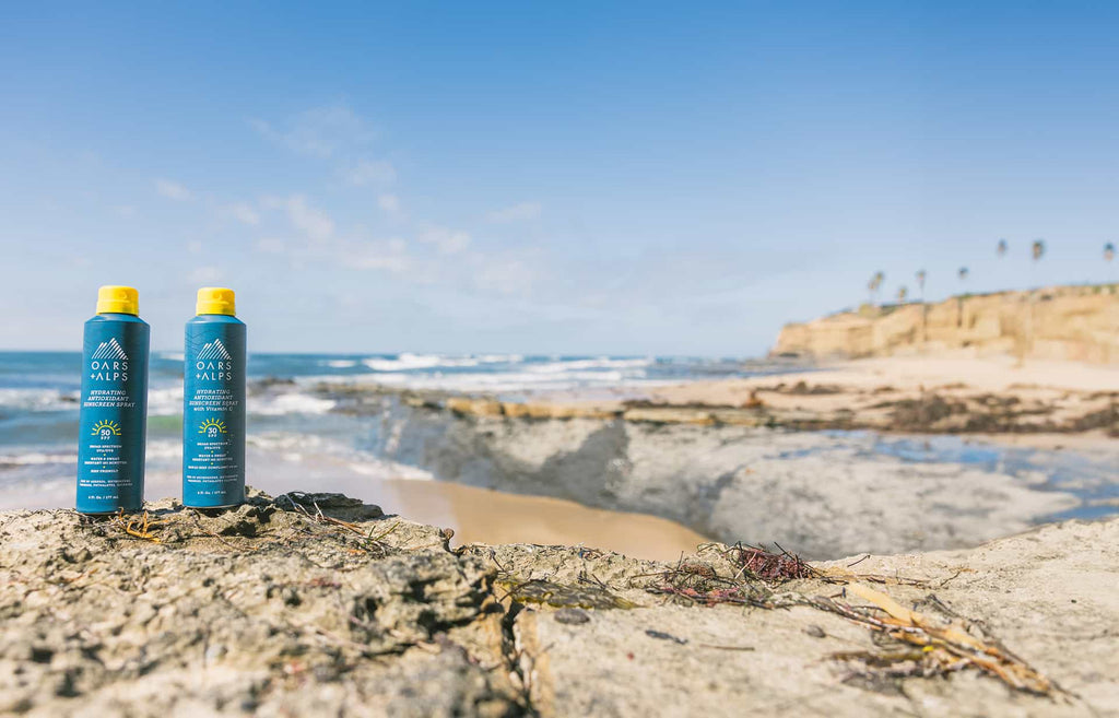 Oars + Alps SPF 50 Sunscreen Spray bottles on a rocky beach coast.